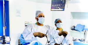 Two surgeons in blue gowns and masks perform surgery with laparoscopic instruments in a bright operating room with monitors nearby.