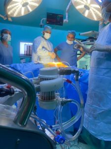 Surgeons in scrubs and masks around an illuminated operating table during surgery in an OR setting with overhead lights nearby.