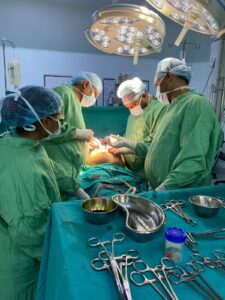 Surgeons in green gowns and masks perform an operation under bright overhead lights, with surgical instruments laid out on a blue draped table.