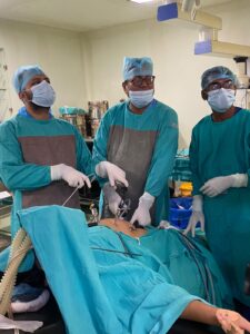 Three surgeons in teal scrubs and masks perform surgery around a patient on an operating table in a sterile OR.