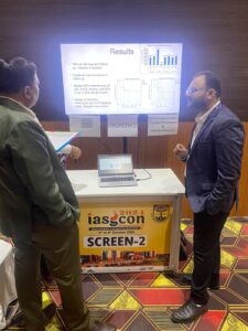 Two men in business attire discuss a poster presentation at an IASCON 2024 booth; a laptop sits on the table.
