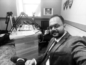 Radio host in a suit and glasses sits in a broadcast studio with microphone and mixing console nearby, taking a selfie in the corner of the room.