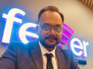 Self-portrait of a man in a blue suit and tie wearing glasses, in front of a bright neon Facebook sign with blue and purple glow.