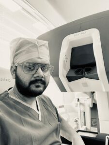 Surgeon in scrubs and a cap posing beside a DaVinci robotic surgery system in the operating room.