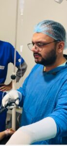 Surgeon in blue scrubs and a cap preparing to perform an operation, with medical equipment and IV stand nearby.