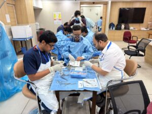 Group of medical trainees in blue gowns and gloves practicing a surgical procedure around a tool-filled tray in a bright training room