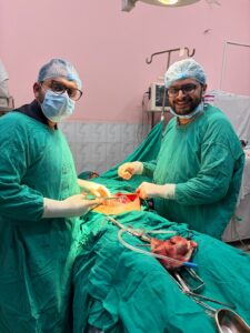 Two surgeons in green scrubs and masks perform surgery at the patient’s side in an operating room, with instruments nearby and a monitor in the background.